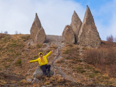 Sarı ceketli ve yüz maskeli bir kadın elleri ayrı bir tepede sivri dişlere benzeyen keskin taş blokların dibinde oturuyor. Eski magma piramitleri
