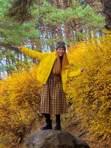 Γελαστή γυναίκα που δείχνει τα δάχτυλα στα πλάγια στέκεται σε μια πέτρα που περιβάλλεται από κίτρινους φθινοπωρινούς θάμνους και ψηλά πεύκα στο πάρκο — Φωτογραφία Αρχείου
