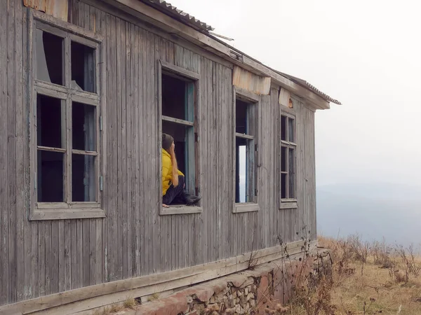 Una chica se sienta en la ventana de una vieja casa abandonada de madera con una niebla mística alrededor — Foto de Stock