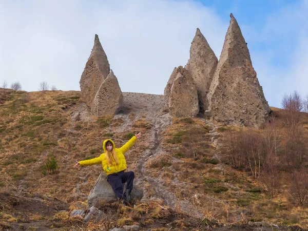 Женщина в жёлтой куртке и маске для лица сидит с раскинутыми руками на холме у подножия острых каменных блоков, напоминающих клыки. Пирамиды магмы — стоковое фото