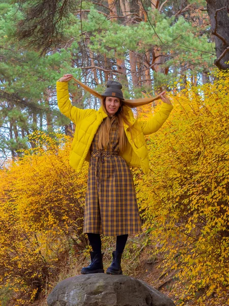 Μια χαρούμενη γυναίκα με μοντέρνα στολή στέκεται κρατώντας τα μαλλιά της σε μια πέτρα σε ένα πάρκο το φθινόπωρο ανάμεσα σε κίτρινους θάμνους. — Φωτογραφία Αρχείου