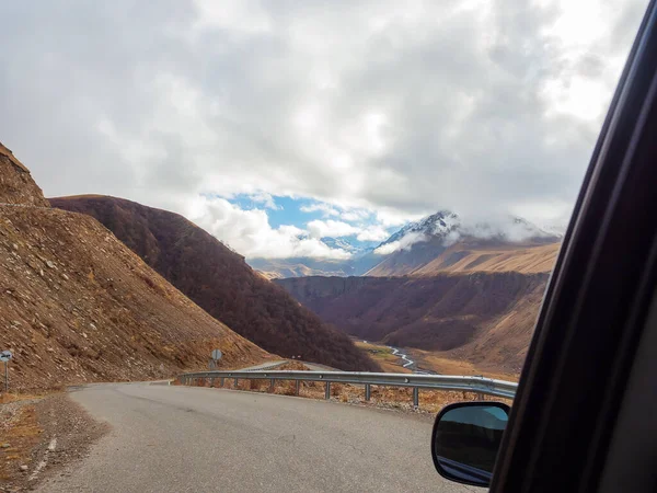 Eine schmale Straße in einer Gebirgsschlucht mit Blick auf den Fluss in der Ferne und Wolken, die auf die Gipfel der Berge herabsteigen. Blick aus dem Autofenster — Stockfoto