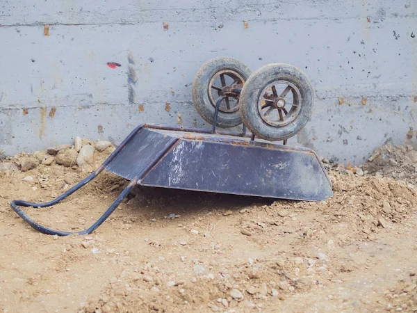 Dirty work cart lies upside down on sand and stones against a concrete wall — Stock Photo, Image