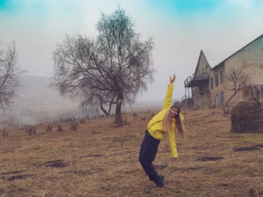 Terk edilmiş bir yerde, samanlıkların arasında, terk edilmiş bir evin arka planına ve sisli sise karşı parmak uçlarında duran bir kadın.