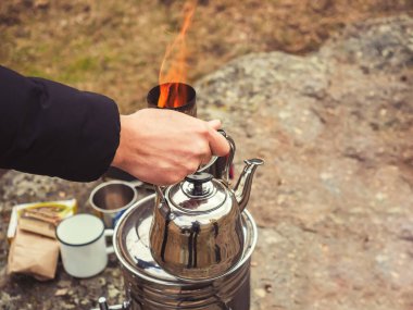 Bir adamın eli krom kaplama bir çaydanlığı semaver bacasından çıkan yanan bir ateşin arka planına bastırır. Bir sonbahar günü doğada çay içmek.