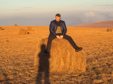 Parlak güneş ışınlarıyla aydınlatılan gülümseyen bir adam sonbahar altın tarlasında samanlıkta oturur..