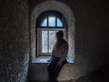 Terk edilmiş eski karanlık bir binada kırık bir pencerenin yanında oturan bir kadın..