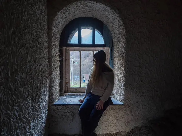 Terk edilmiş eski karanlık bir binada kırık bir pencerenin yanında oturan bir kadın..