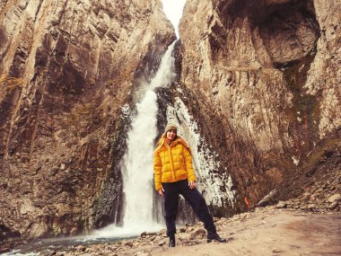 Gülümseyen bir kadın, Kafkas dağlarındaki kayalık bir vadiden akan buz gibi bir şelalenin arka planında duruyor.