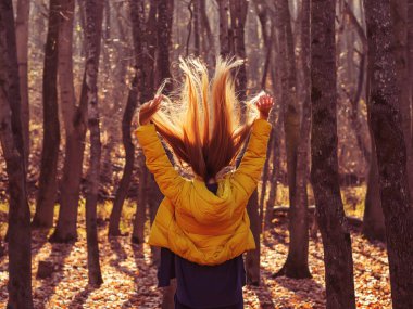 Bir kadın, bir sonbahar günü düşen yaprakların arasında bir sonbahar parkında dikilirken, uzun saçlarını güneşin arkasına atar.