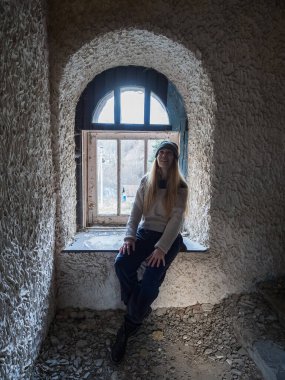 Terk edilmiş eski karanlık bir binada kırık bir pencerenin yanında oturan bir kadın. Eski harabelerde yolculuk