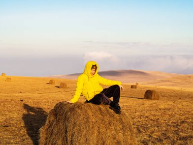Parlak sarı ceketli bir kadın sonbahar tarlasında, parlak güneş ışığıyla aydınlatılmış bir samanlıkta oturuyor.