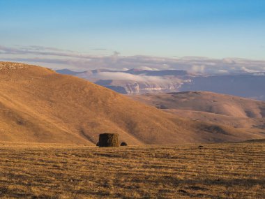 Mavi gökyüzü ve alçak bulutların altında yüksek bir dağ vadisinde yuvarlanmış bir saman yığını. Kafkasların doğası.