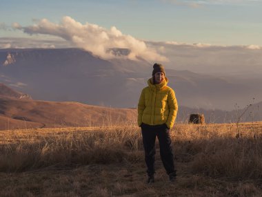 Bir kadın, sonbaharda, dağın tepesinde, bulutlarla kaplı bir platoya karşı, Kafkas dağ vadisiyle çevrili altın çimenlerde duruyor..