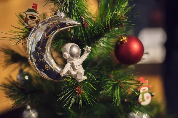 A tiny figurine of an astronaut sits on a shiny silvery moon. Christmas tree decoration. Holiday poster.
