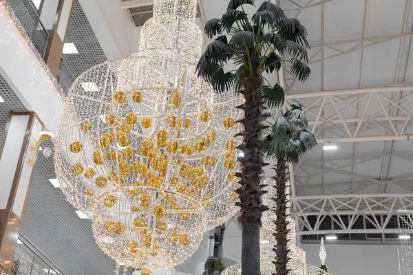 En stor ljuskrona festligt dekorerad med kransar och gyllene nyårskulor hänger i hallen i köpcentret på nivån för höga palmer. Stockbild