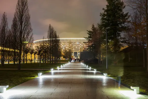 Parque nocturno con un sendero peatonal iluminado que conduce a un gran edificio luminoso semicircular. Imagen De Stock