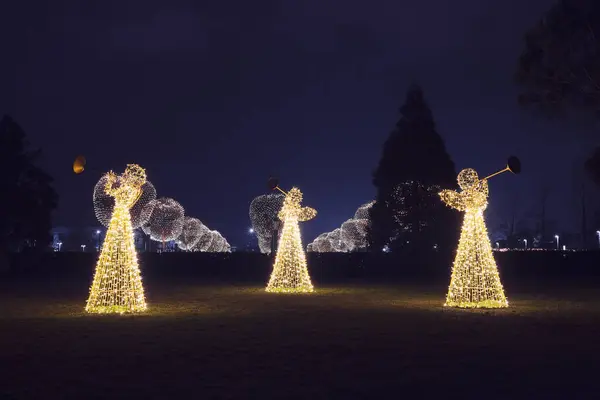 Three Christmas angels from luminous garlands illuminate the park walking area against the backdrop of round luminous trees at night. Festive decor