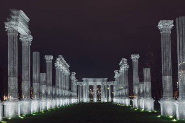 Rusya, Krasnodar 07.01.2022. Beyaz çerçeveli parlak sütun, gece parkında karanlık bir gökyüzünün altında yeşil bir ara sokak süslüyor.