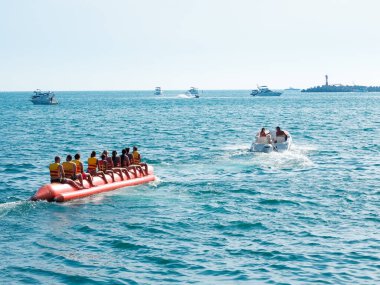 Bir grup insan yaz günü denizde şişme muz teknesine biniyor. Deniz eğlencesi. Güney tatili.