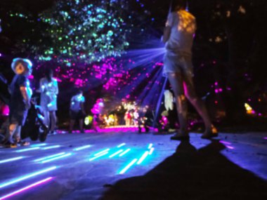 Parlak projeksiyonlarla aydınlatılmış insanlar geceleri parkta yürürler. Lazer gösterisi. Işık ve gölgelerin uzun pozlama oyunu. Bulanık resim