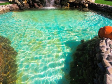 Güneş altında parlayan turkuaz suyla dekoratif yaz göleti ve taş bir duvardan aşağı akan şelale.
