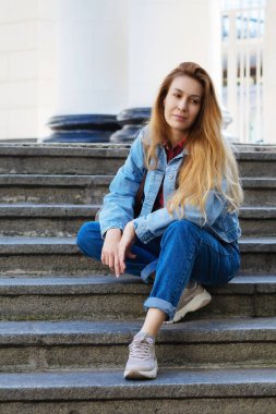Kot pantolon giymiş, açık renk saçlı bir kadın sokak merdivenlerinde oturuyor. Şehir klasik mimarisi. Modern moda.