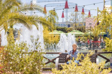 Rusya, Soçi 14.05.2022. Bir kadın, bir eğlence parkında fıskiyeyle gölün arka planında bankta oturuyor. Bahar güneşinin tadını çıkarıyorum. Güney turizmi.