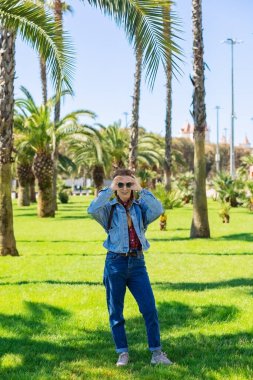 Palmiye ağaçlarının yanında yeşil çimlerde dikilen bir kadın parmakları açık gözlüklerine tutunuyor. South Park 'ta aylak aylak dolaşmak