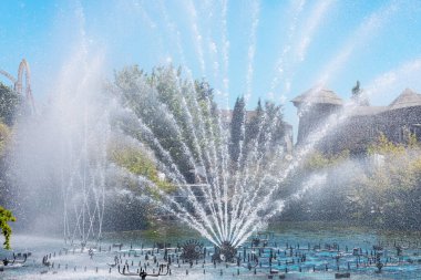 Güneşli bir günde, bir lunaparkta bulunan sığ mavi göletin ortasındaki fan şeklinde bir çeşmenin güçlü akıntıları.