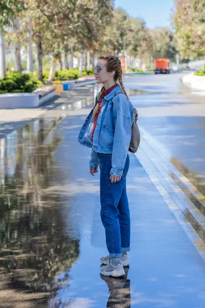 A woman in jeans clothes stands on wet alley in a spacious spring park. Walk after the rain. Youth modern fashion.
