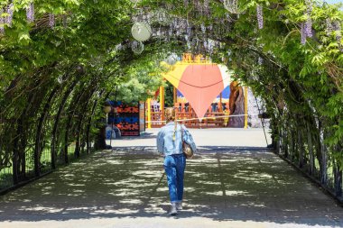 Bir kadın, çok renkli bir lunaparkta akasya ile sarmalanmış bir kemerin altında dar bir sokakta yürüyor.