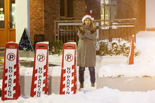 Woman showing forbidding gesture on street. woman near sign in winter. No parking sign in winter. Woman in santa hat at evening in winter yard