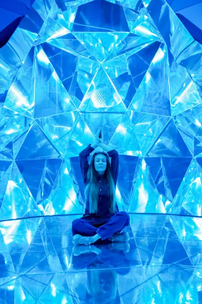 Nilüfer pozisyonundaki bir kadın ellerini başının üstüne yapıştırmış, parlak desenli bir ayna mozaik odasında oturuyor.