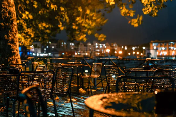City cafe terrace near the river in the rainy autumn evening in the lantern light. Raindrops on outdoor metal furniture. Urban fall concept.Selective focus. — Stock Photo, Image