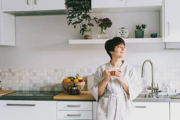 Femme heureuse dans le peignoir tenant une tasse de thé frais dans sa cuisine blanche moderne. Bonjour, concept. Ambiance positive. Concentration sélective. Espace de copie. — Photo
