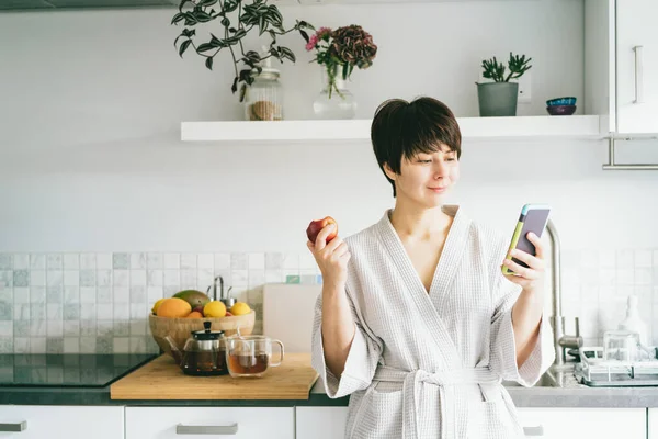 Eine lächelnde Frau im Bademantel blickt auf ihr Handy und isst einen Apfel, während sie in der mordenden Küche steht. Morgens Gewohnheiten und Rituale. Online-Sucht, Konzept zur Tagesplanung. — Stockfoto