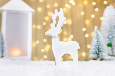 Beyaz ahşap geyik oyuncağı, fener ve karların üzerinde küçük Noel ağaçları olan Noel masalı kompozisyonu. Mutlu Noeller ve mutlu yıllar kartı. Seçici odak.