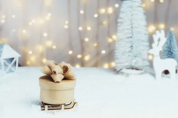 Composición de cuento de hadas de Navidad de un trineo de madera con regalo ecológico en la nieve con árbol de Navidad, ciervos y luces cálidas fondo bokeh. Feliz Navidad y feliz año nuevo tarjeta. Enfoque selectivo. — Foto de Stock