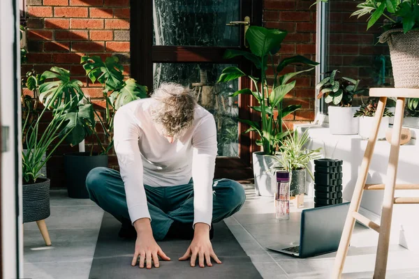 Hombre joven sentado en la estera, haciendo estiramiento de la posición de yoga en acogedora habitación de estilo loft moderno llena de plantas caseras en la mañana soleada. Práctica de yoga en línea. Un estilo de vida saludable. Inicio entrenamiento en línea. — Foto de Stock