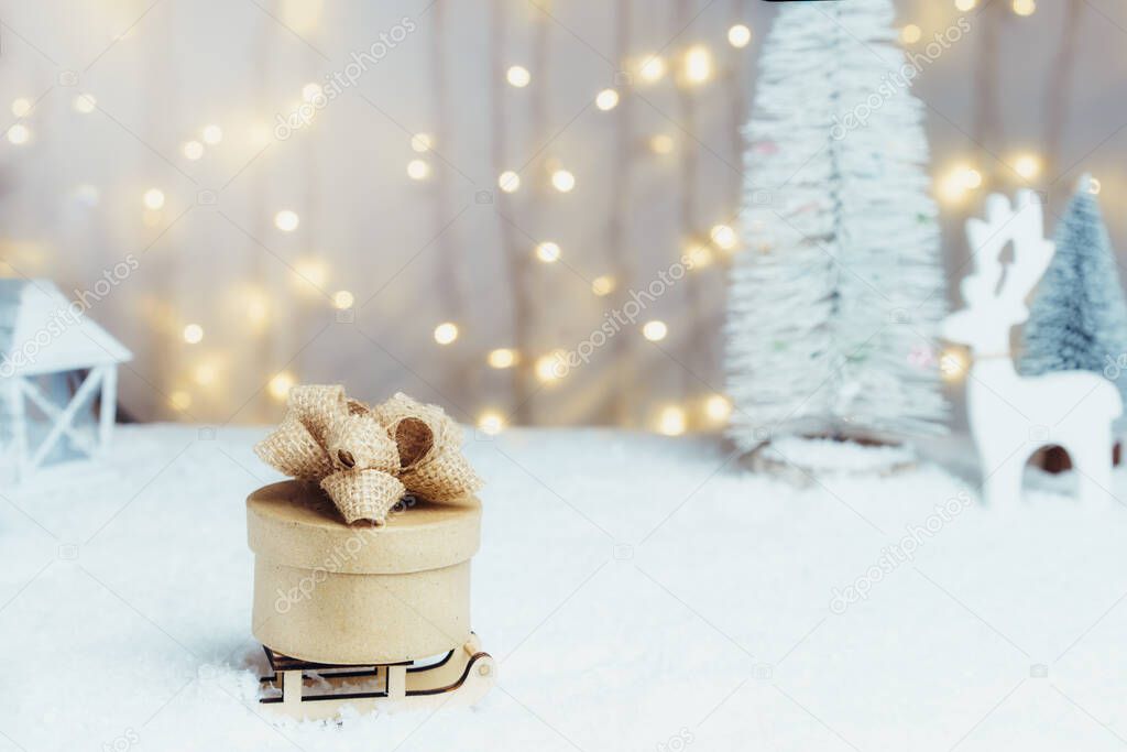 Christmas fairy tale composition of a wooden sleigh with eco-friendly gift on the snow with Christmas tree, deer and warm lights bokeh background. Merry Xmas and Happy new year card. Selective focus.