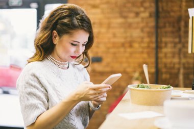 Kafede yemek yerken akıllı telefonuna bakan konsantre bir kadın. Uzak çalışma, sosyal ağlar kayıyor, sohbet ediyor veya notlar alıyor.