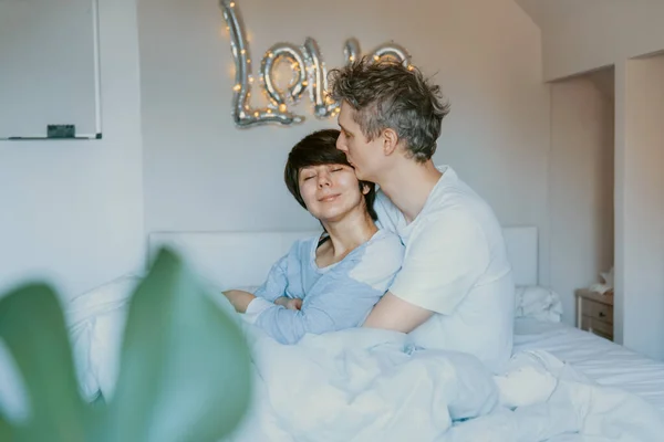 Kochająca para w piżamie bawiąca się w łóżku w sypialni ze słowem Miłość na tle ściany. Przytulają się i śmieją. Szczęśliwych rodzinnych poranków w domu. Walentynki. Skupienie selektywne. — Zdjęcie stockowe