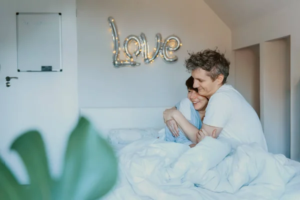 Älskade par i pyjamas har roligt i sängen i sovrummet med ord Kärlek på väggen bakgrund. De omfamnar och skrattar. Glad familjemorgon hemma. Alla hjärtans dag. Selektiv inriktning. — Stockfoto