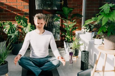 Paspasın üzerinde oturan genç adam, güneşli bir sabahta ev bitkileriyle dolu, rahat, modern bir çatı katında yoga pozisyonu alıyor. Çevrimiçi sınıf yoga pratiği. Sağlıklı yaşam tarzı, meditasyon. Evde çevrimiçi çalışma.