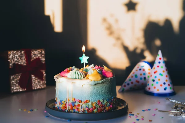 Färgglada födelsedagstårta med strössel och stjärnformade ljus, festliga mössor och presentask på en mörk vägg bakgrund med skuggor. Selektivt fokus, kopieringsutrymme. — Stockfoto