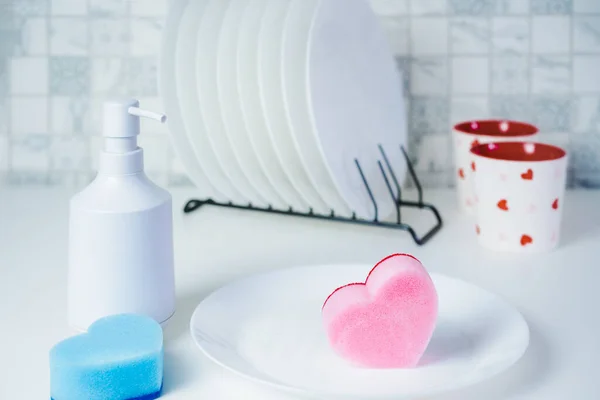 Esponjas en forma de corazón, jabón lavavajillas en el recipiente blanco y platos limpios y tazas en la mesa de la cocina. Composición moderna de los utensilios de cocina. Suministros de limpieza bio orgánicos. Copiar espacio. Enfoque selectivo. — Foto de Stock