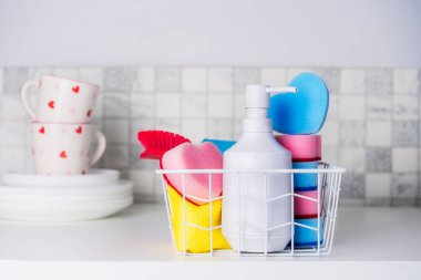 Oda servisi konsepti. Tabak sabunu, kalp şeklinde süngerler, sepetteki fırça ve haplar mutfak masasındaki temiz tabaklar ve bardaklar. Modern mutfak malzemeleri kompozisyonu. Seçici odak.