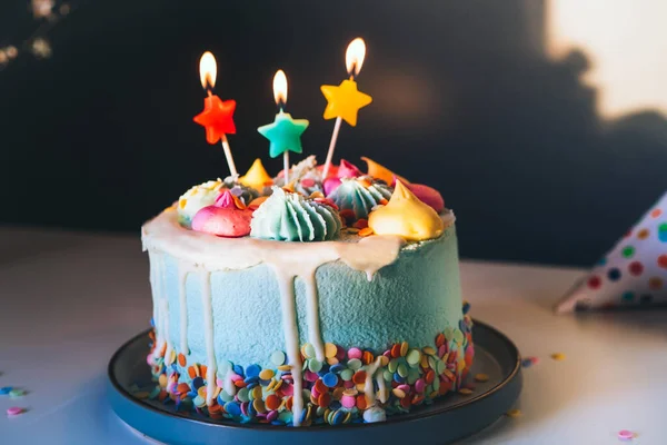 Colorful birthday cake with sprinkles and burning star shaped candles on a dark wall background with shadows. Festive birthday celebration, party. Selective focus, copy space — Stock Photo, Image