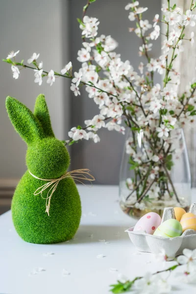 Grüner Osterhase mit blühenden Blumen auf dem Ast in der Vase und farbigen Ostereiern im Gefäß auf dem weißen Tisch. Frohe Ostern Frühling vertikale Karte. Selektiver Fokus. Kopierraum. — Stockfoto